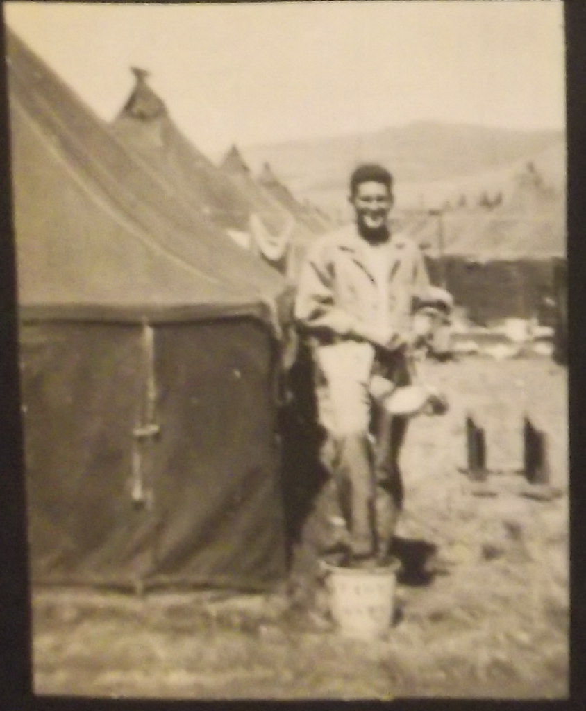 A sepia image of a young White man standing outside of a row of tents.