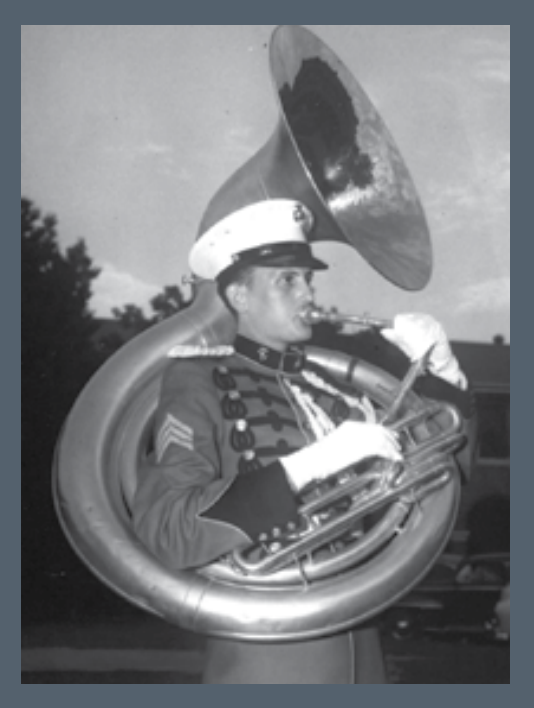 A black and white image of a young White man playing the tuba dressed in uniform.