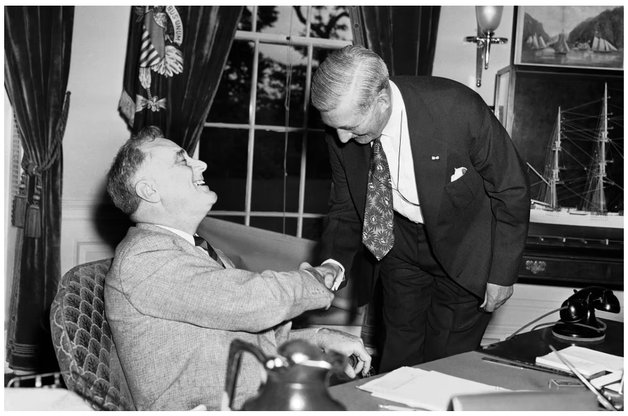 A black and white image of two older White men shaking hands. One is sitting behind a desk and the other is standing and leaning over.