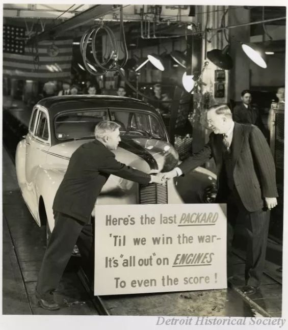 A black and white photo of a car on the assembly line. In front of the car are two older White men shaking hands in front of a sign that reads. “Here’s the last Packard ‘Til we win the war - It’s ‘all out’ on Engines to even the score!”