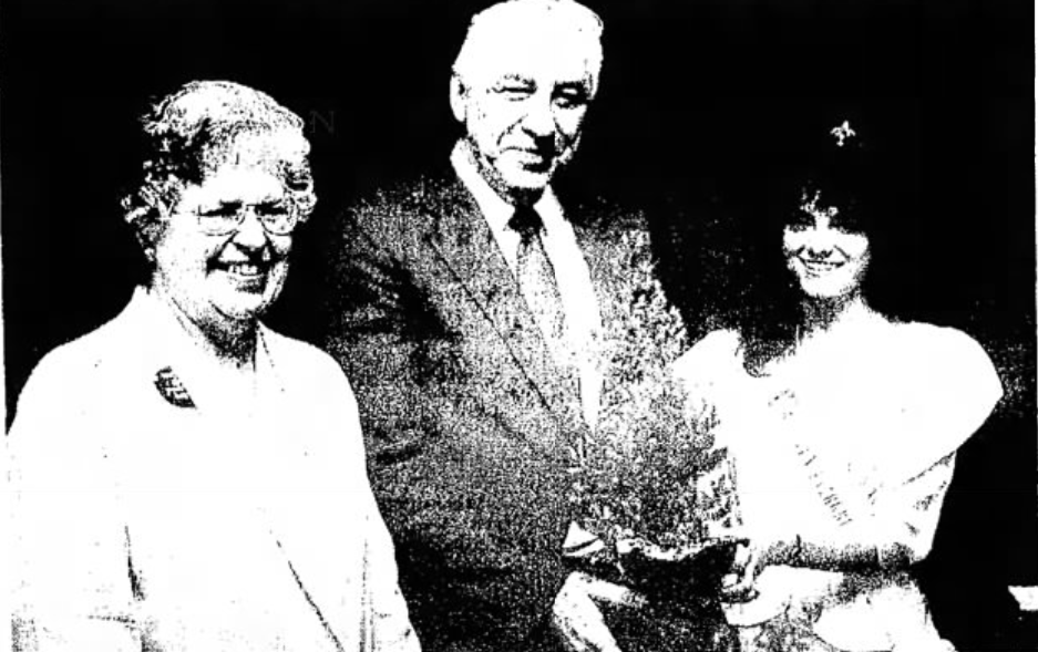 A black and white image of an older White man in a suit and tie standing in between an older White woman and a younger White woman.