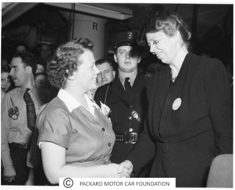 A black and white image of an older White woman shaking hands with a younger White woman in a crowded room with a policeman standing guard.