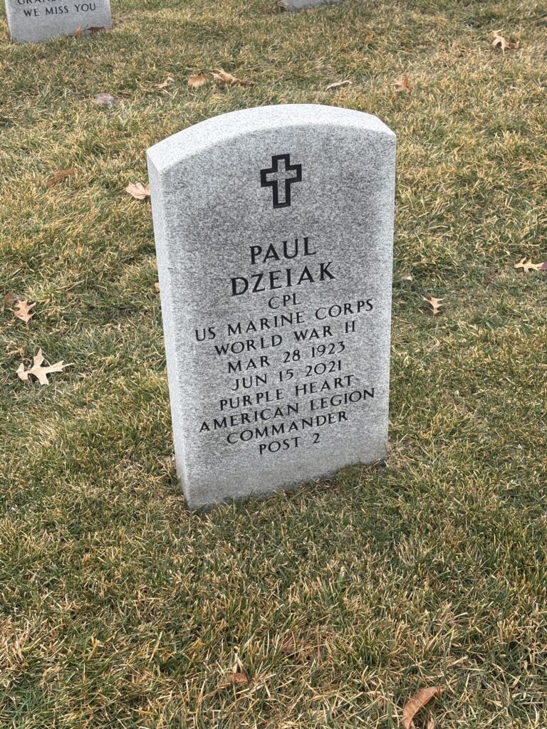 A gray marble arched headstone with a cross on top reads, “Paul Dzeiak CPL US Marine Corps World War II Mar 28 1923 Jun 15 2021 Purple Heart American Legion Commander Post 2.”