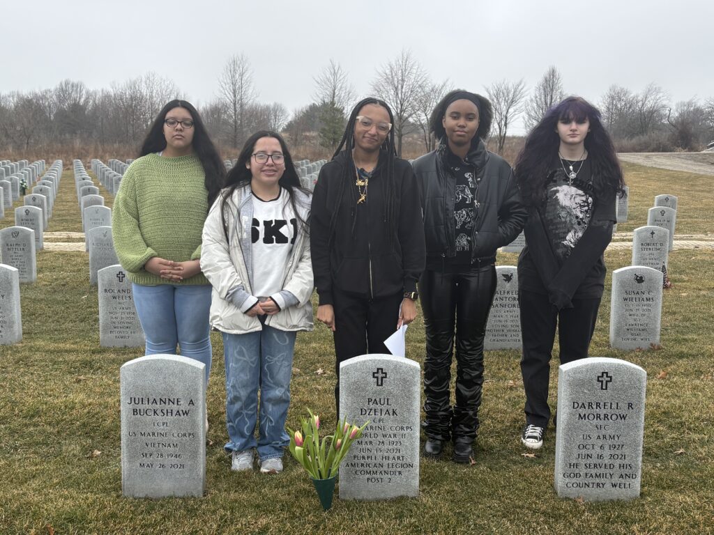 A color photo of five students (all girls) standing behind Dzeiak’s headstone. They have placed tulips next to his grave.