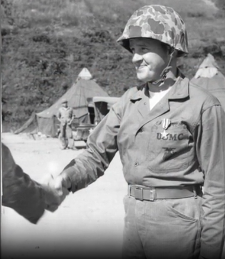 A black and white image of a young White soldier in uniform shaking hands with an unseen person. They are outside, in the field.