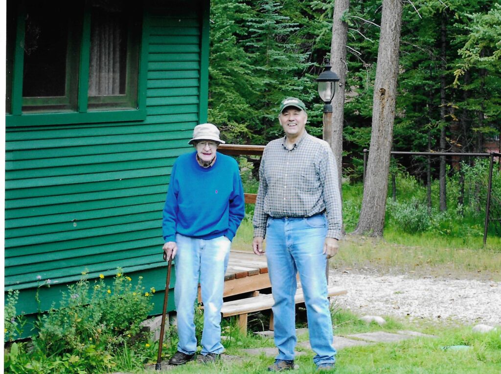 A color image of an elderly White man and a middle age White man standing outside a green cabin.