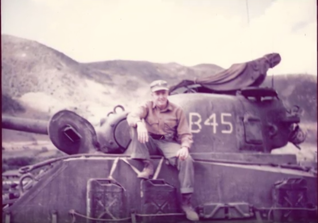 A color image of a young White man sitting on the outside of a tank. He is posing for the image.
