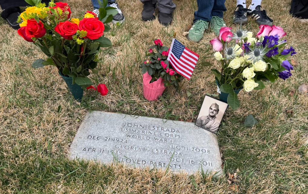 A color image of a headstone with flowers, a photo, and a flag