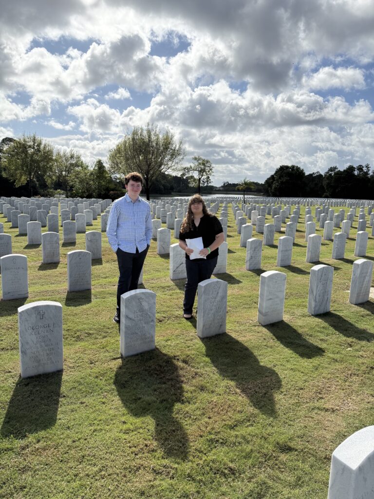 A color photo of two students standing behind a row of headstones in a cemetery.