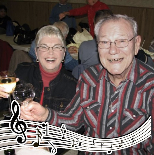 A color photo of an elderly White man and woman sitting at a table. They are dressed casually, sitting at a table, and toasting. 