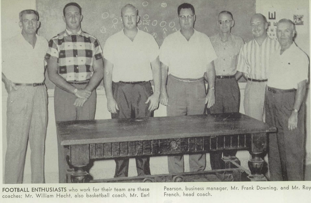 A black and white image of seven White men  standing behind a big wooden table inside a classroom. They are all dressed in button-up shirts and slacks.