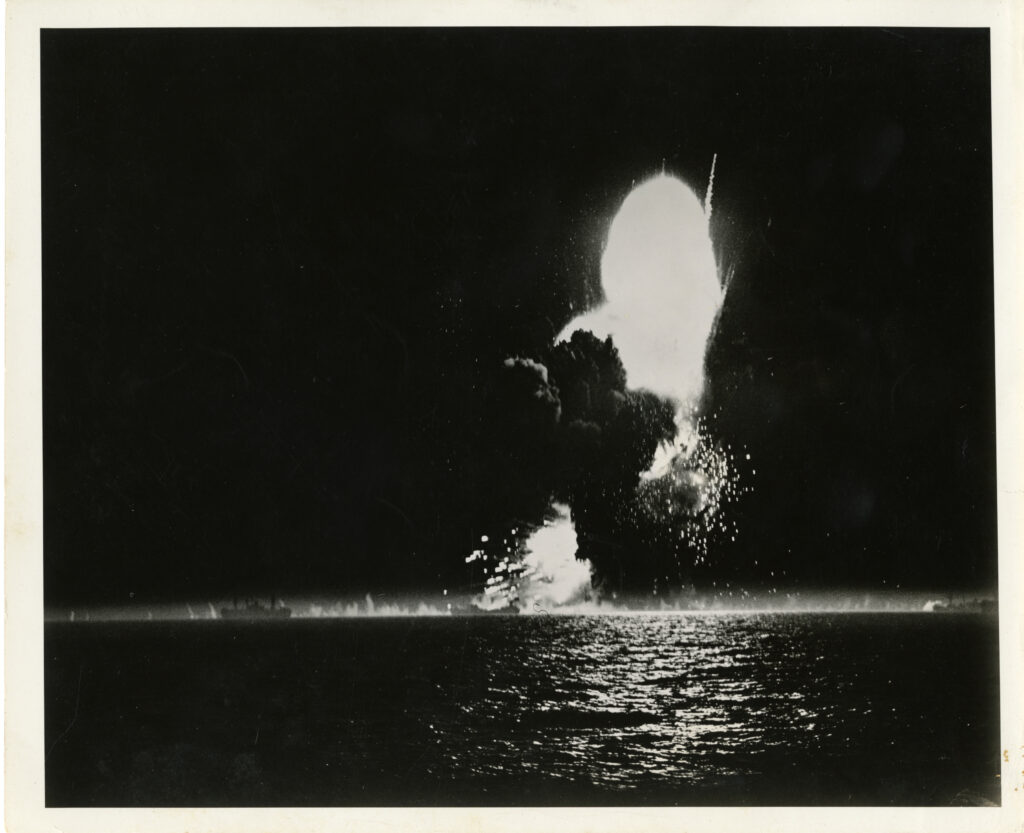 A black and white photograph of a body of water with an explosion in the distance. The fire is rising into the sky, and water is splashing around it, where debris is hitting the water.