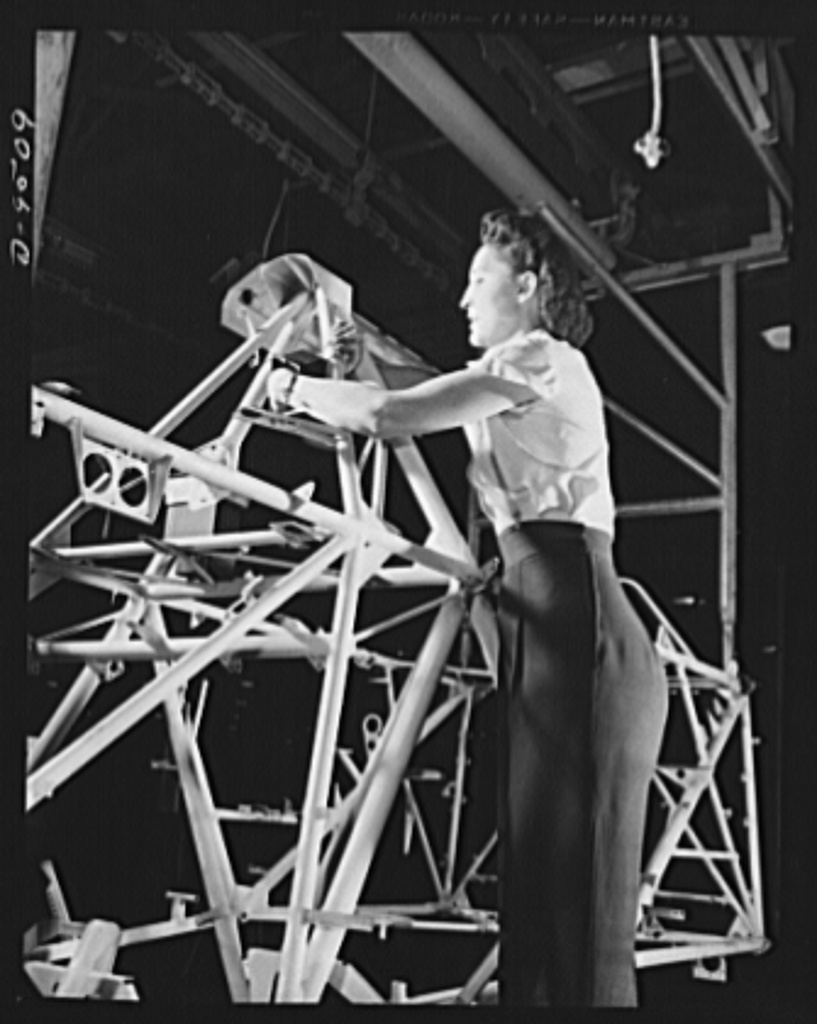 A black and white image of a woman working on the top part of an airplane frame.