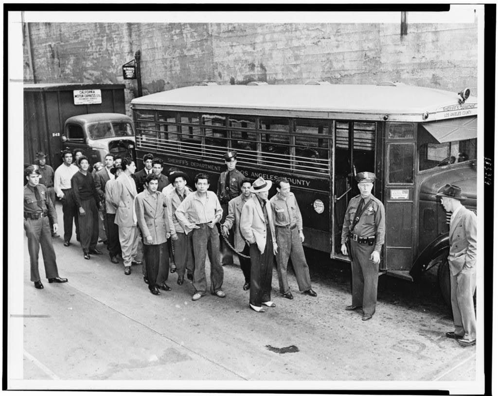 A black and white photograph of several young men in zoot suits outside a police transport bus. A few police officers are guarding them.
