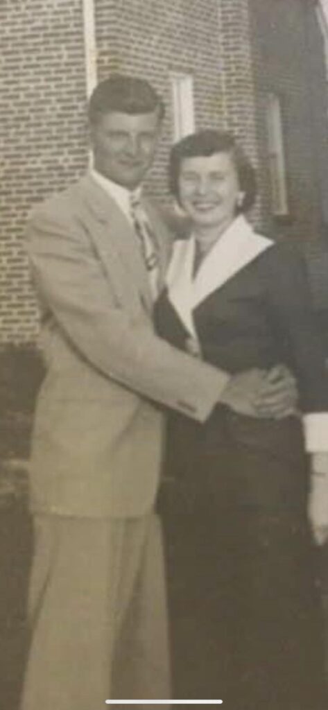 A black and white image of a young White couple holding each other while looking at the camera. The man is in a suit and tie. The woman is in a pantsuit.