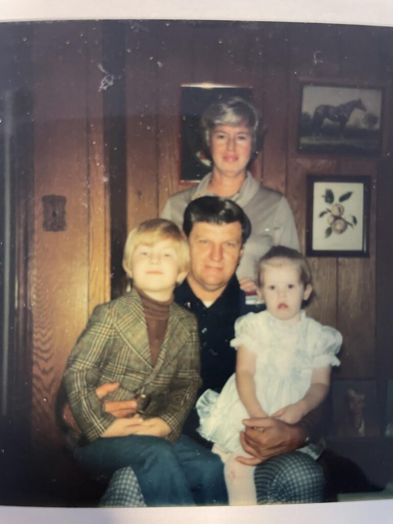 A color photo of an older White man sitting in a chair holding a young boy and a toddler-aged girl on either knee. Behind him stand a White woman. They are inside a home.