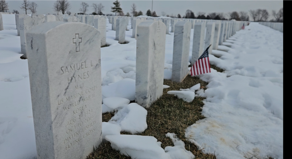 A color photo of several arched white marble graves in a row. In the foreground is one engraved with a cross at the top and the words “Samuel L. Hynes MAJ U.S. Marine Corps World War II Korea Aug 29, 1924 Oct 10 2019 Distinguished Flying Cross.”