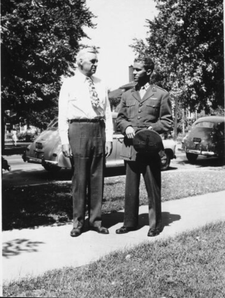 A black and white image of a young White man in uniform standing next to an older White man on a neighborhood street.