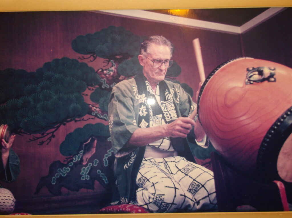 A color image of an elderly White man in traditional Japanese clothing playing a drum on his knees.