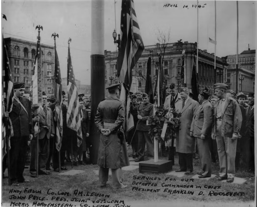 A black and white image of many people in uniform gathered around a flagpole with the flag lowered.