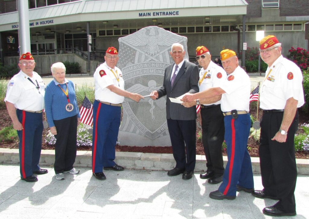 A color image of several older people in uniform standing outside a building. One is handing another a check.