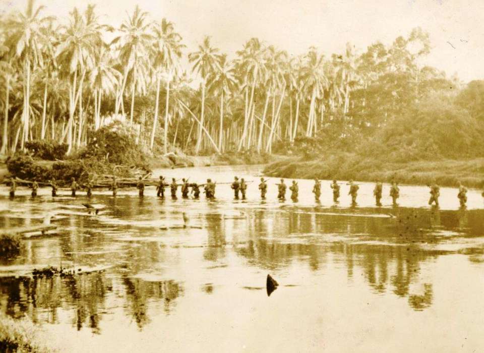A sepia image of a few dozen Marines crossing a knee-high river in the middle of a jungle.