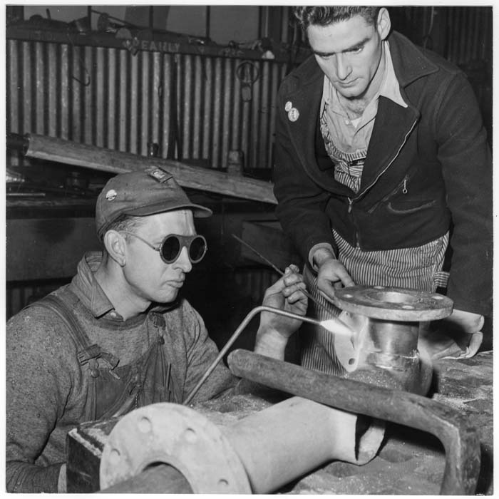 A black and white image of two White men welding some type of pipe in a factory.