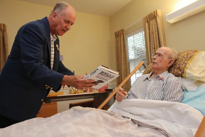 A color image of an elderly White man in a hospital bed talking with an older White male who is holding a book.