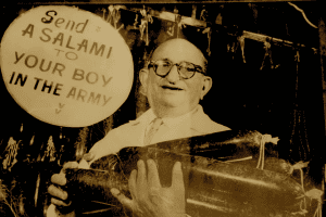 A black and white image of an older White man in glasses with his arms full of salami. Behind him there is a sign with the slogan.