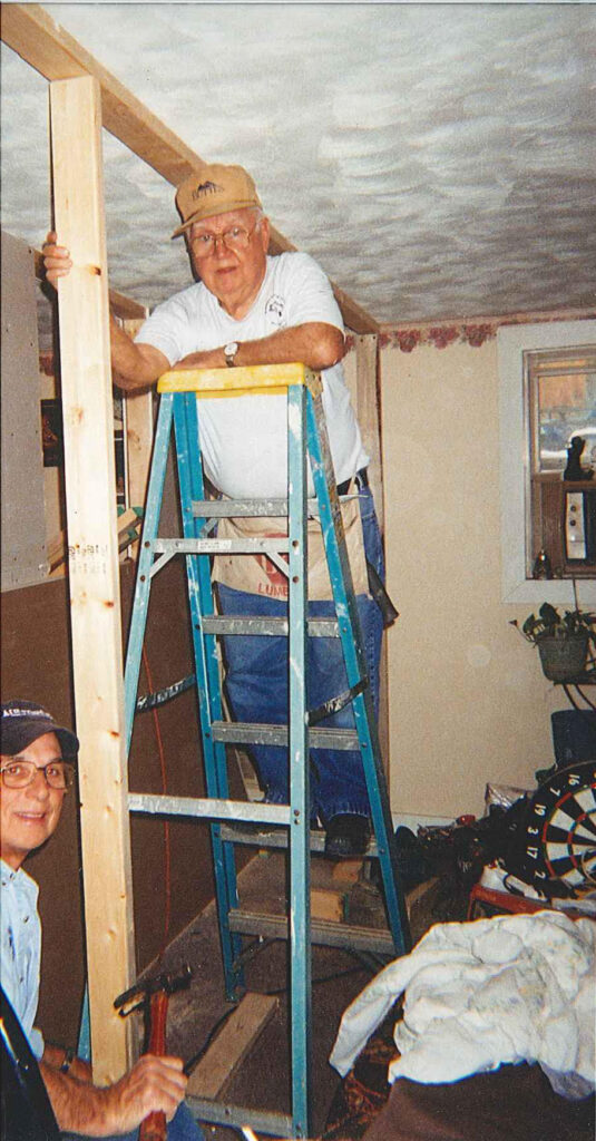 A color photo of an elderly White man on a ladder holding up a 2x4 inside a house.