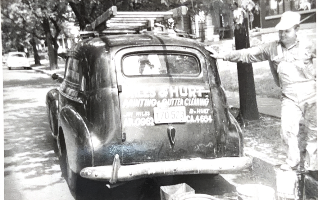 A black and white photo of a younger White man leaning up against an old-style truck
