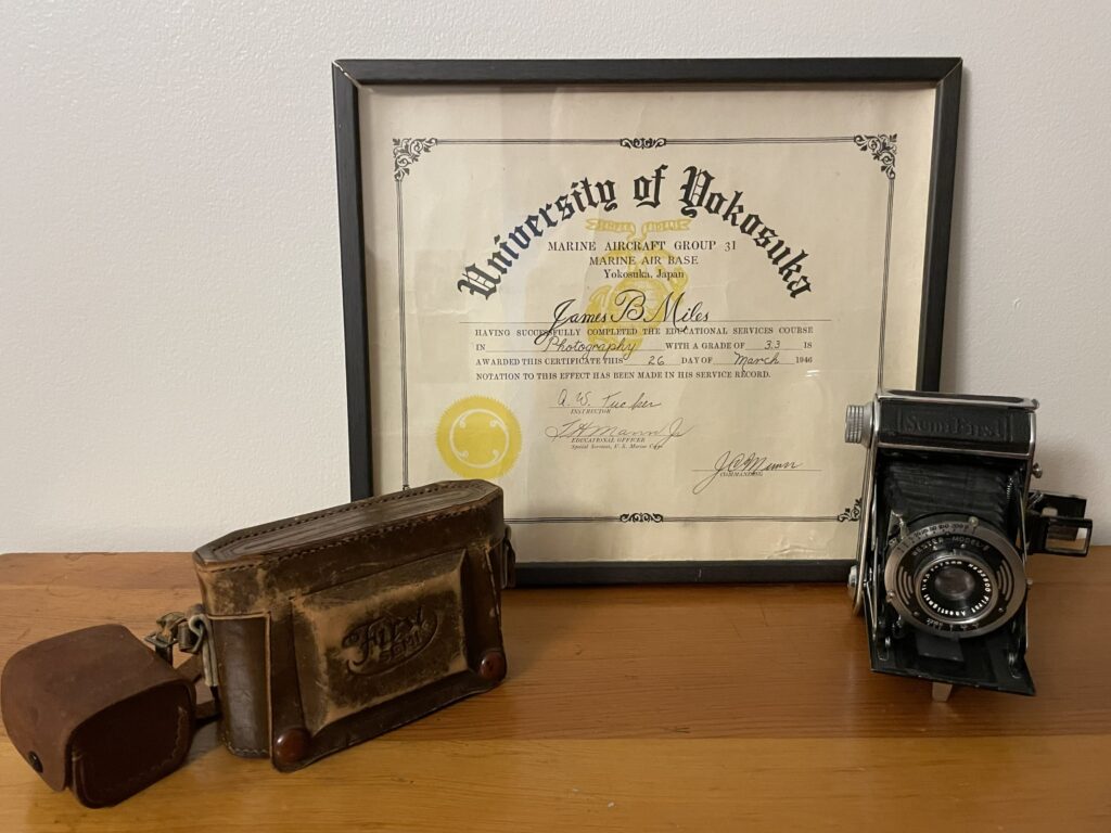A color photo of an old style camera, leather bag, and a framed certificate.