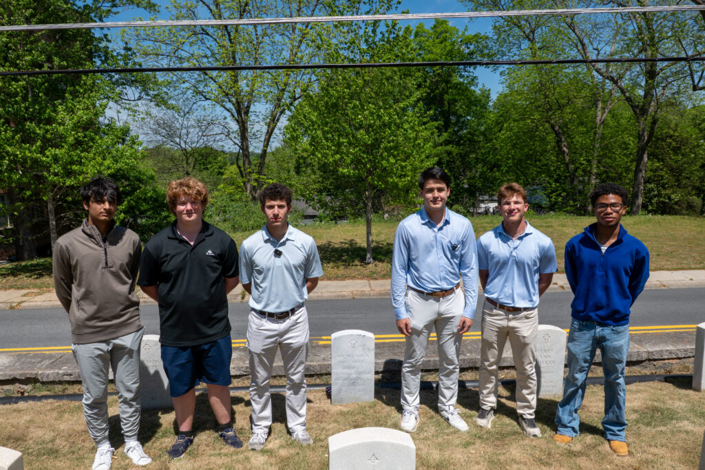 A color photograph of six young men standing next to a headstone in a cemetery.