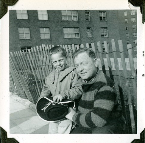 A black and white photo of a young White boy standing with an older White man who is kneeling down to the boy. They are standing in front of a fence outside of what appears to be a large apartment building.