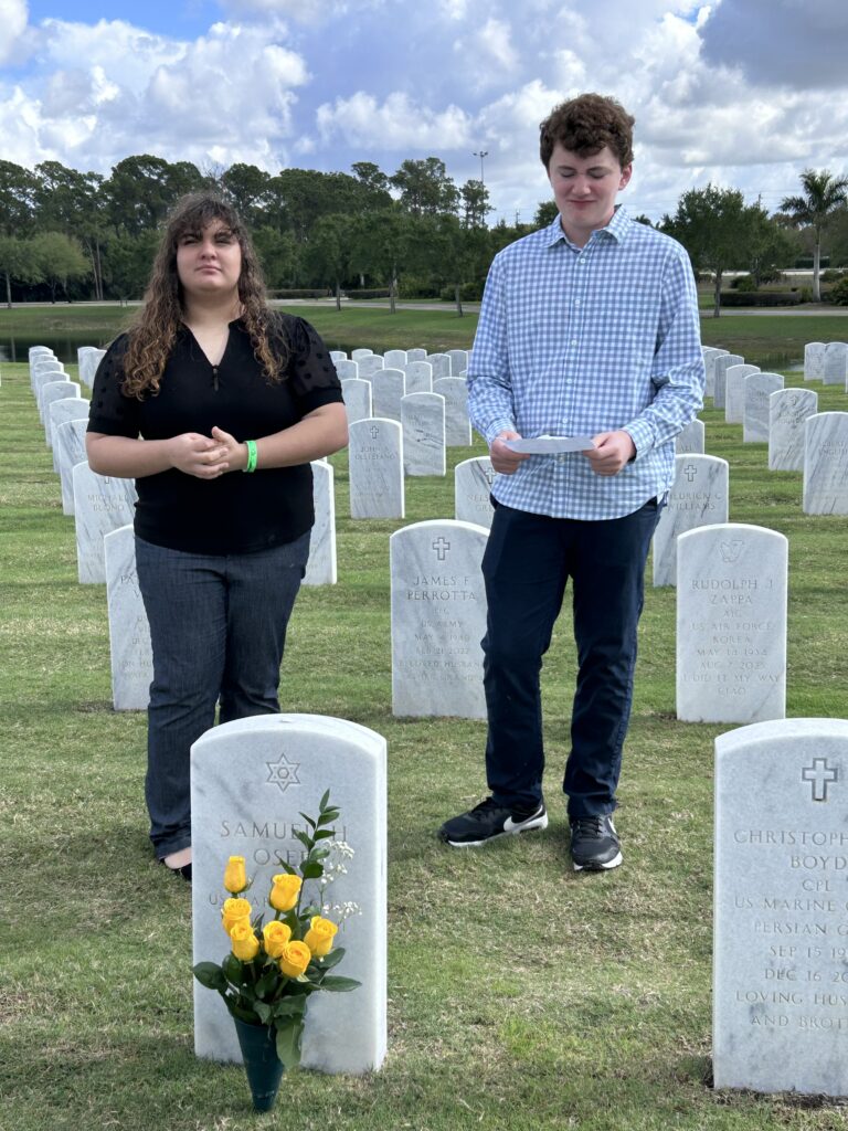 A young man and woman stand behind a headstone with flowers in front.