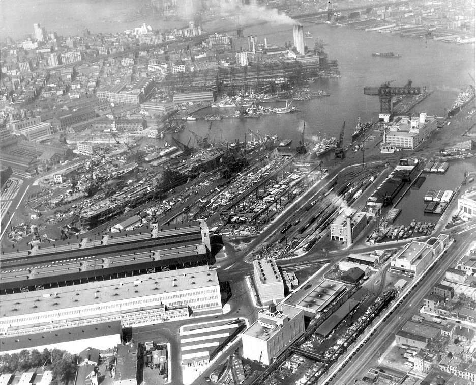 A black and white aerial photo of a large Navy shipyard with multiple boats in production.