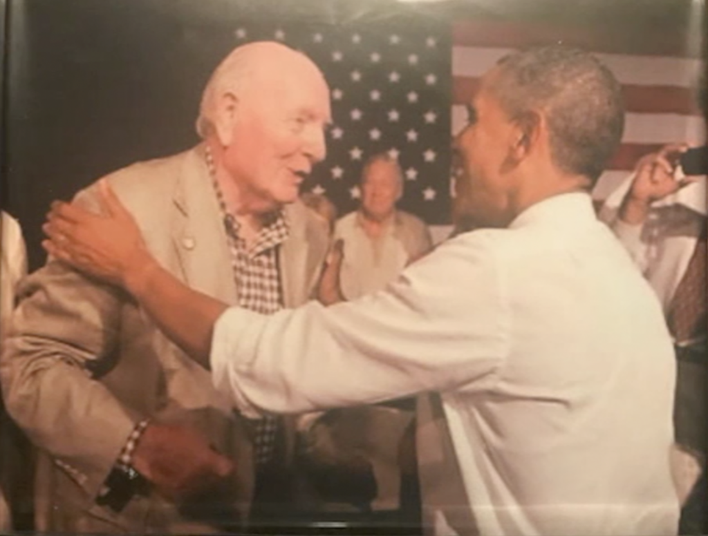 A color image of a middle age Black man in a white dress shirt shaking hands with an older White man in a suit jacket.