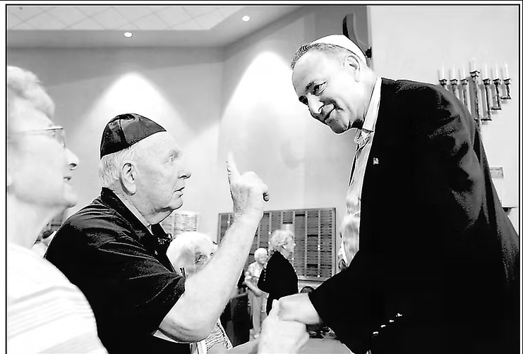 Black and white image of an older White man in a suit and yalmika shaking hands with an elderly White woman in a church. Another elderly White man is talking to him.