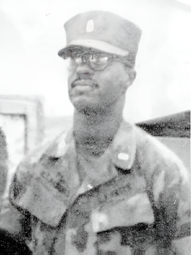 A black and white photograph of a young Black man in a jungle utility uniform. He appears to be standing at attention.