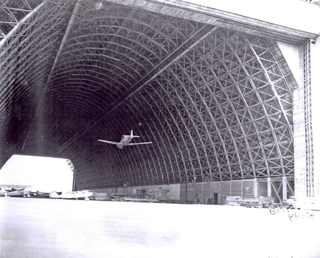 A black and white photo of a large airplane hangar made of wood. There are several planes parked and one is flying through it inside.