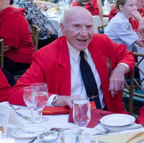 A color photo of an elderly Whie man sitting at a dinner table in a red blazer with a white button-up shirt and black tie.