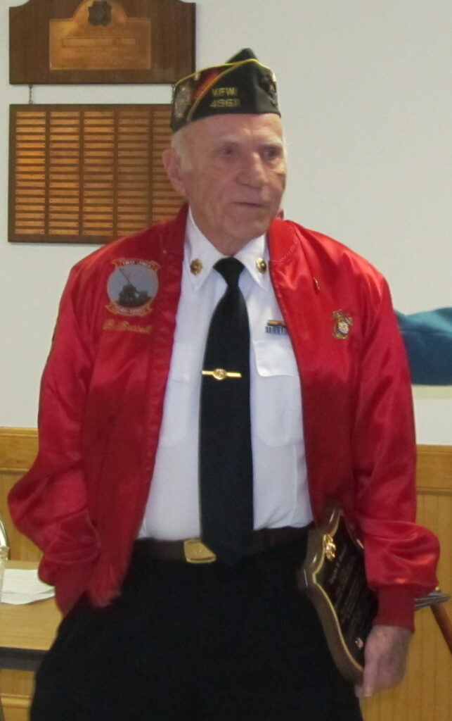 A color image of an elderly White man in a dress shirt and tie with a red coat. He is holding a plaque.