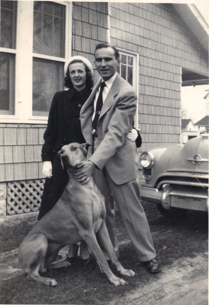 A black and white photo of a younger White couple standing outside a house and car with a Great Dane.