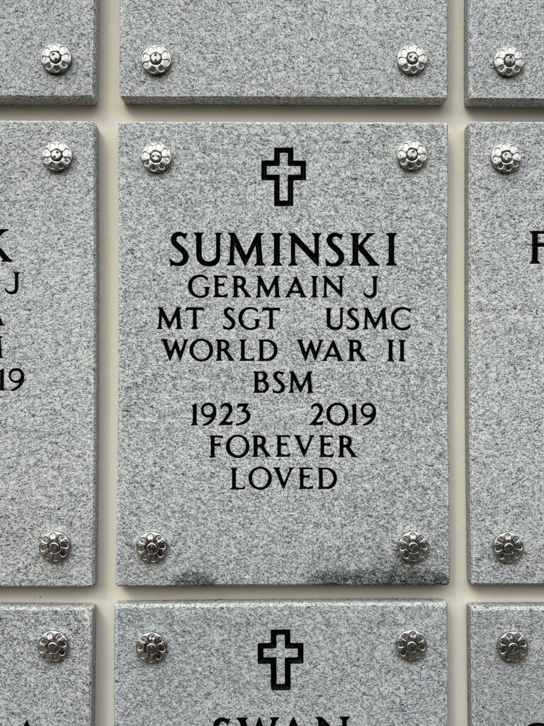 A grey, rectangular stone engraved with a cross at the top and the words, “Suminski Germain J MT SGT USMC World War II BSM 1923 2019 Forever Loved.”