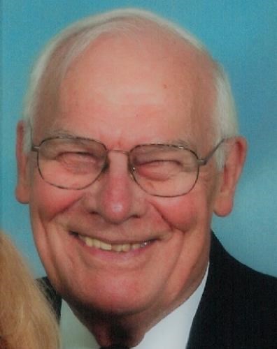 A color headshot of an elderly White man in glasses. He is wearing a suit and tie and smiling at the camera.