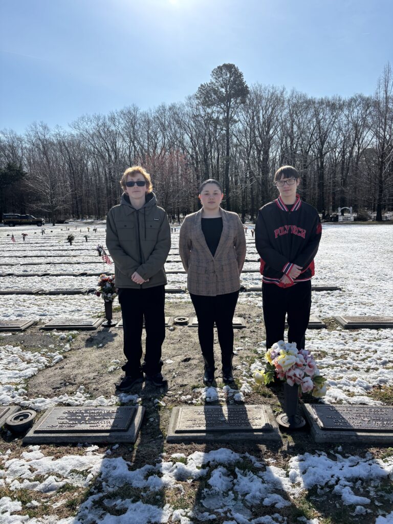 Three students (two boys and one girl) stand behind flat gravestones.