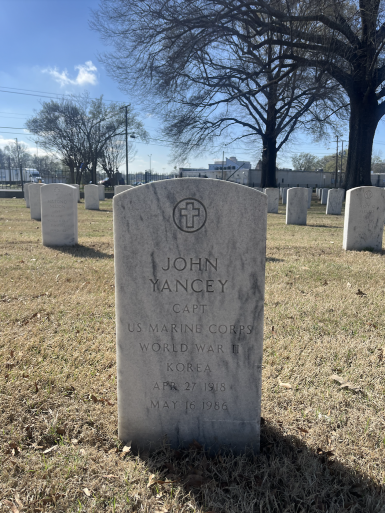 An arched white marble headstone with a cross at the top is engraved with “John Yancey Capt US Marine Corps World War II Korea Apr 27 1918 May 16 1986.”