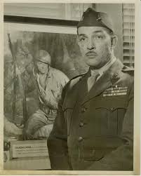 A black and white image of a middle-aged Black man standing in uniform in front of a painting of a Marine in battle.