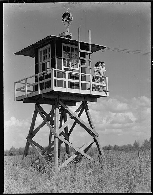 A black and white image of a guard tower in an open field. Two young people are seen out on the balcony.