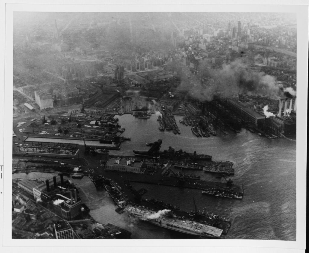 A black and white aerial image of many ships being built on the shores of a massive city.